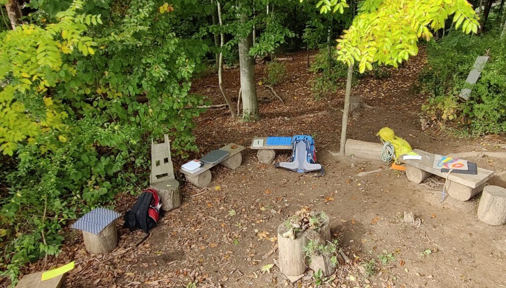 The photograph shows a forest clearing with seating arranged in a circle, backpacks and writing material. The image represents workshops and coaching for individuals in the greater Stuttgart and Karlsruhe area.